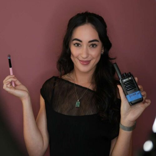 Emmalani Howe smiling behind the scenes of “Barefoot to Hills,” holding a makeup brush and production walkie-talkie in a cinematic studio setting.